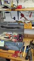 Gray steel toolbox open showing contents including cordless drill, caulking guns, clamps, and assorted hand tools.