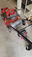 Photo showing the side view of the red Troy-Bilt lawn mower with string trimmer laid over it and a red gas can beside it