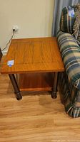 Square wood end table next to plaid couch on wooden floor.