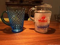 Two glass jugs side by side on wooden surface: one blue with knobby texture and one clear with red and blue nautical ship design.