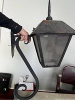 Side view showing the wrought iron bracket with decorative scroll design and the square mounting plate. Visible surface rust on edges of the lantern's metal frame.