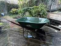 Side view of green Garant metal wheelbarrow with red wood handles and black tire on a wooden deck outdoors.