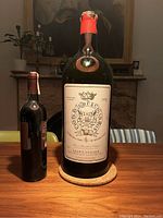 Photo of large 5 litre green glass wine bottle with wax cap, positioned next to a smaller standard wine bottle for scale, on a table indoors.