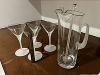 Set of four clear martini glasses with frosted stems and bases next to clear glass pitcher with frosted handle and glass stopper, on a wooden surface