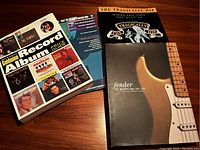 Four music related books on a wooden surface: Goldmine Record Album Price Guide, Power Studies TAB (with visible cassette), The Tragically Hip Original Rock Charts The Best Of music score book, and Fender The Golden Age 1946-1970 history coffee table book.