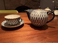 Tea pot and matching teacup with saucer on wooden table under warm lighting. Blue and white lattice pattern with gold accents visible.