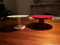 Two ceramic cake stands on wood table, one white (8 inch diameter) and one red with gold rim (10 inch diameter), both with scalloped edges and pedestal bases.