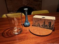 Photo of three vases: one clear glass vase, one blue and black speckled ceramic narrow-neck vase, and one rectangular ceramic vase with village scene on cork mat.
