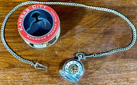 The pocket watch with attached chain and red and black round tin container labeled 'Kansas City Railroad Pocket Watch' displayed on wooden surface.