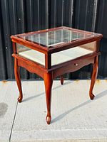 Overall view of the wooden side table with glass display case and Queen Anne style cabriole legs on a concrete surface outside.