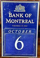 Full frontal view of the vintage Bank of Montreal dark blue perpetual calendar metal sign showing the logo, text, and calendar set to October 6.