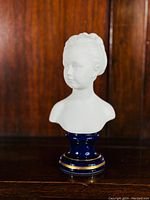 Front view of bisque porcelain bust of a girl on cobalt blue and gold glazed plinth, wooden background.