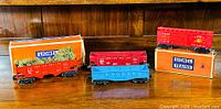 Five vintage Lionel O Gauge freight train cars shown with three original boxes. The cars include red, blue, and orange colors with detailed markings. Boxes show Lionel branding and a freight yard set label.