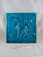 Full view of the antique blue ceramic tile showing embossed scene with two men indoors near a fire and various objects.