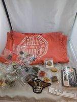 Overview of orange-red Harley Davidson tank top with white logo spread with pins, patches, and sealed playing cards in front
