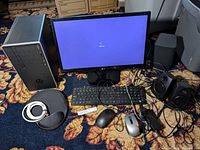 HP Pavilion desktop tower, LG LED monitor powered on showing Windows 10 lock screen, two wired mice, black wired keyboard, black mouse pad, black router, and black external speakers arranged on floral carpet.