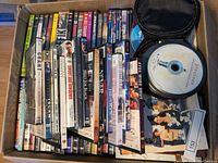 Top view of a large box filled with multiple unsealed DVD cases arranged standing upright, some titles visible including 'Grant', 'Traffic', 'Triple X', and others, plus a small black carrying case holding loose DVDs.