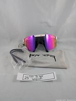 Front view of sunglasses with purple mirrored lens, white frame with pink splatter, soft pouch, additional black temple arms in plastic.