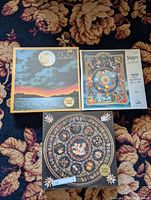 Three sealed vintage puzzles stacked on carpet: F.X. Schmid 605 piece moonlit landscape, Suns Out Tarot 1000 piece by Ciro Marchetti, and F.X. Schmid Florentine mosaic 132 piece puzzle.