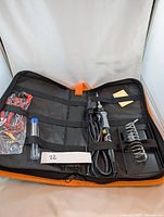 Soldering kit opened in a black and orange fabric case showing soldering iron, solder wire spool, some small accessories including sandpaper and cleaning tools.
