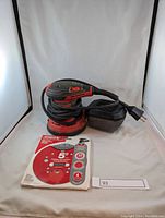 Front view of the Milwaukee sander with power cord wrapped around it beside a pack of Diablo sanding discs.