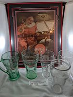 Framed 1951 Coca-Cola Christmas ad with 5 collectible Coca-Cola glass cups displayed in front, showing different green and clear glass styles.