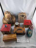 Full lot of seven vintage wooden coin banks arranged on white background, showing carved coconut monkey, houses, barrel, box, and clog.