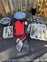 Kitchen items on black cloth including pottery bowls, casserole dish, red oven mitts, metal cooking utensils, and a tray of silverware.