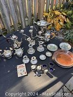 Full view of all the silver plated items arranged on a black cloth outdoors, including teapot, sugar bowl, creamer, candle sticks, small dishes and other pieces.
