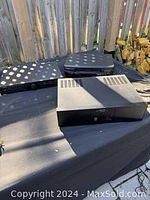 Three electronic units outdoors on black surface: Rotel amplifiers and Panasonic VHS player