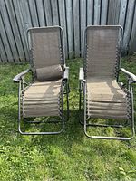 Front view of two brown gravity chairs placed on grass near a wooden fence. Both chairs have metal frames and brown mesh fabric seating. One chair has a lumbar support pillow attached.