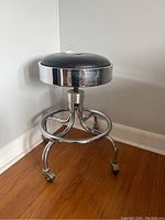 Side and angled view of adjustable metal stool showing black padded round seat and chrome frame with four legs and caster wheels.