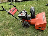 Side view of orange two-stage snowblower showing engine, black chute, and tire with chains.