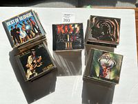Stack of CDs including Best of Blondie, Kiss Alive III, Van Halen, and several others spread in six small stacks on a white surface under sunlight.