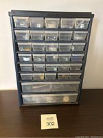 Front view of black plastic organizer with multiple small transparent drawers filled with assorted nuts, bolts, screws, washers, and other small hardware items.