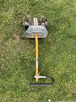 Top-down view of the Ryobi cultivator attachment placed on grass, showing the 4-metal tine cultivator head with Ryobi branding and the yellow handle with black grip loop at the end.