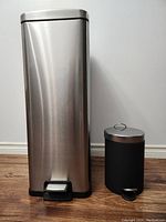 Both garbage cans side by side on wood floor against white wall to show size comparison and condition.