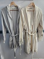 Photo of two hung bathrobes, one beige and one white with gray stripes, both with waffle weave and fringed edges.