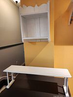 Two white wooden shelves shown in a home setting against a yellow wall. Bottom shelf is long and narrow on floor, hanging shelf mounted above with scalloped edge.