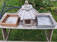 View of the three bird feeders displayed on a wooden bench outdoors: a large wooden hexagonal bird feeder in center, two tray feeders to the left and right, one light wood and one metal tray feeder, showing mesh base inside trays.