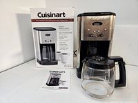 Full shot of Cuisinart 12-cup coffee maker with glass carafe and original packaging and manual in background