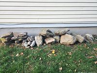 Long row of approximately 40 assorted stones varying from flat flagstones to larger rounded landscaping stones, placed on grass against a white house foundation wall.