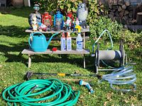 Wide photo showing arrangement of gardening tools and items on lawn and benches including watering can, garden gnomes, lanterns, hoses, and hose reel