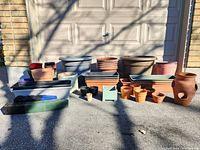 Lot overview showing multiple terracotta, ceramic, and plastic planters and pots arranged on pavement in front of a garage door, varying sizes and colors.