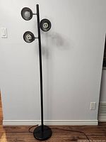 Full view of the black standing three-light lamp against a plain wall, showing base and pole with three lights.