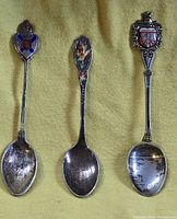 Three silver souvenir spoons displayed front side, showing enamel crests and bowl of spoons.