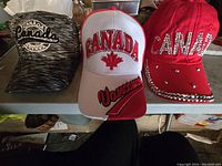 Three Canada themed ball caps: black and gray cap with embroidered patch, white and red cap with 'Canada' and maple leaf embroidery, and a red rhinestone embellished cap.