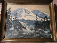 Framed painting showing a trapper's cabin with mountains in the background.