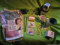 Wide view of all assorted kitchen and gift items arranged on green background including apron, thermos, coasters, bottle openers, mugs, rice cube mold, glass sauce dispensers, and chopsticks.