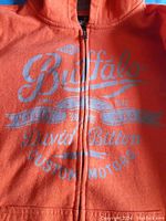 Close-up of the Buffalo David Bitton printed logo on front of the orange zippered hoodie.